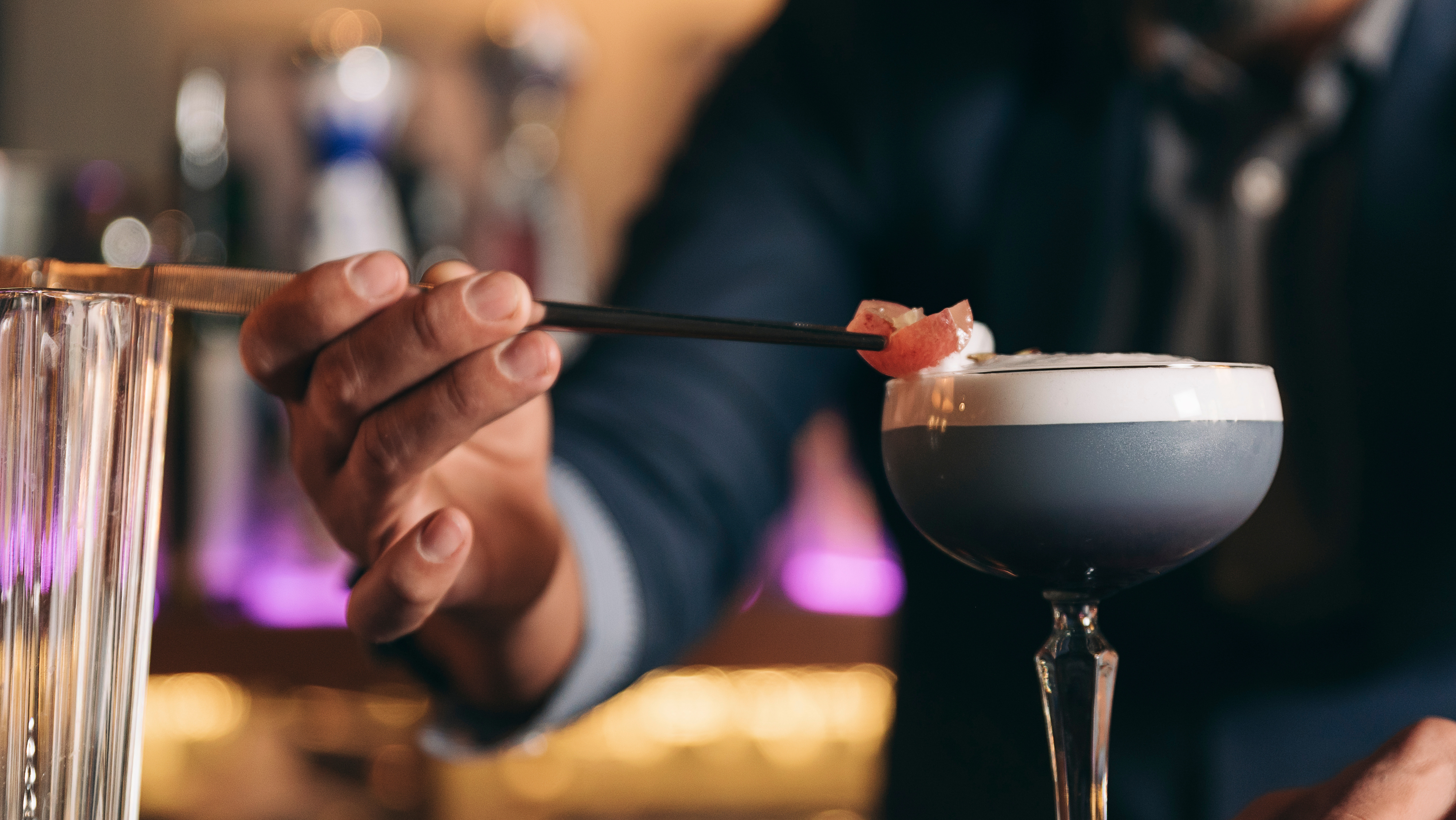 A bartender skillfully places a garnish atop a cocktail using golden bar tweezers.